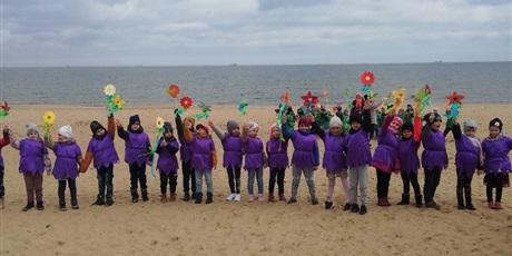 Powiększ grafikę: Na zdjęciu grupa dzieci w wieku przedszkolnym w fioletowych strojach z kwiatkami na plaży