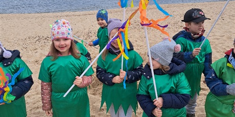 Powiększ grafikę: Na zdjęciu grupa dzieci w wieku przedszkolnym w strojach zielonych z kwiatkami na plaży witająca wiosnę