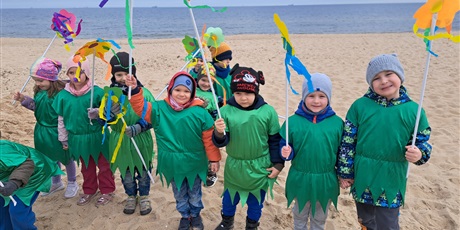 Powiększ grafikę: Na zdjęciu grupa dzieci w wieku przedszkolnym w strojach zielonych z kwiatkami na plaży witająca wiosnę