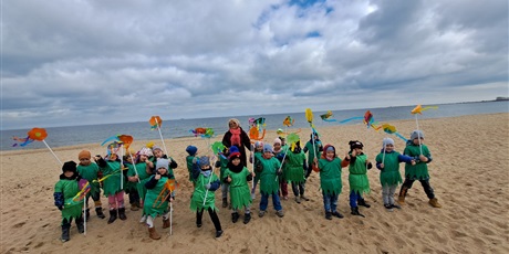 Powiększ grafikę: Na zdjęciu grupa dzieci w wieku przedszkolnym w strojach zielonych z kwiatkami na plaży witająca wiosnę