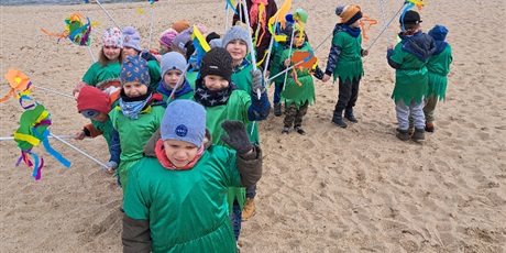 Powiększ grafikę: Na zdjęciu grupa dzieci w wieku przedszkolnym w strojach zielonych z kwiatkami na plaży witająca wiosnę