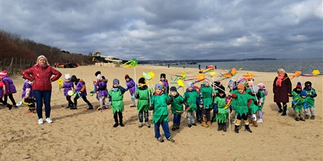 Powiększ grafikę: Na zdjęciu grupa dzieci w wieku przedszkolnym w strojach zielonych z kwiatkami na plaży witająca wiosnę