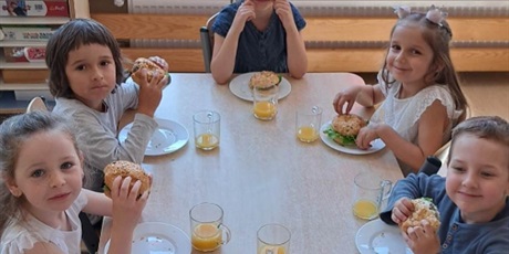 Powiększ grafikę: Na zdjęciu grupa dzieci w wieku przedszkolnym jedząca hamburgery i pijąca sok pomarańczowy.