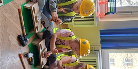 Powiększ grafikę: Na zdjęciu grupa dzieci w wieku przedszkolnym w żółtycha kaskach i kamizelkach odblaskowych podczas warszatów stolarskich.